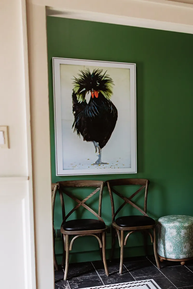 A framed painting of a black chicken hangs on a green wall above two wooden chairs and a patterned ottoman, evoking the cozy charm of a French restaurant.