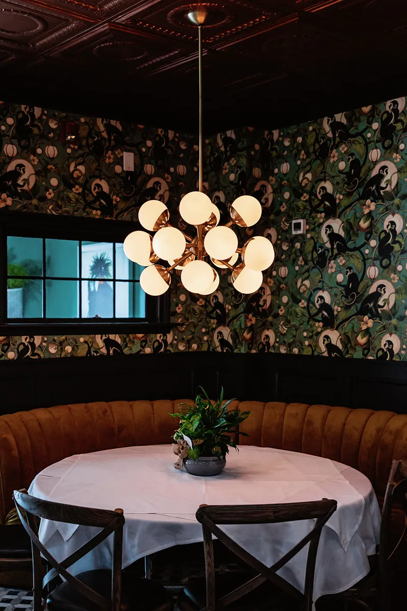 A round table with a white tablecloth and a potted plant centerpiece sits in a corner booth under a modern chandelier, evoking the cozy elegance of a French restaurant, with patterned wallpaper and a window in the background.