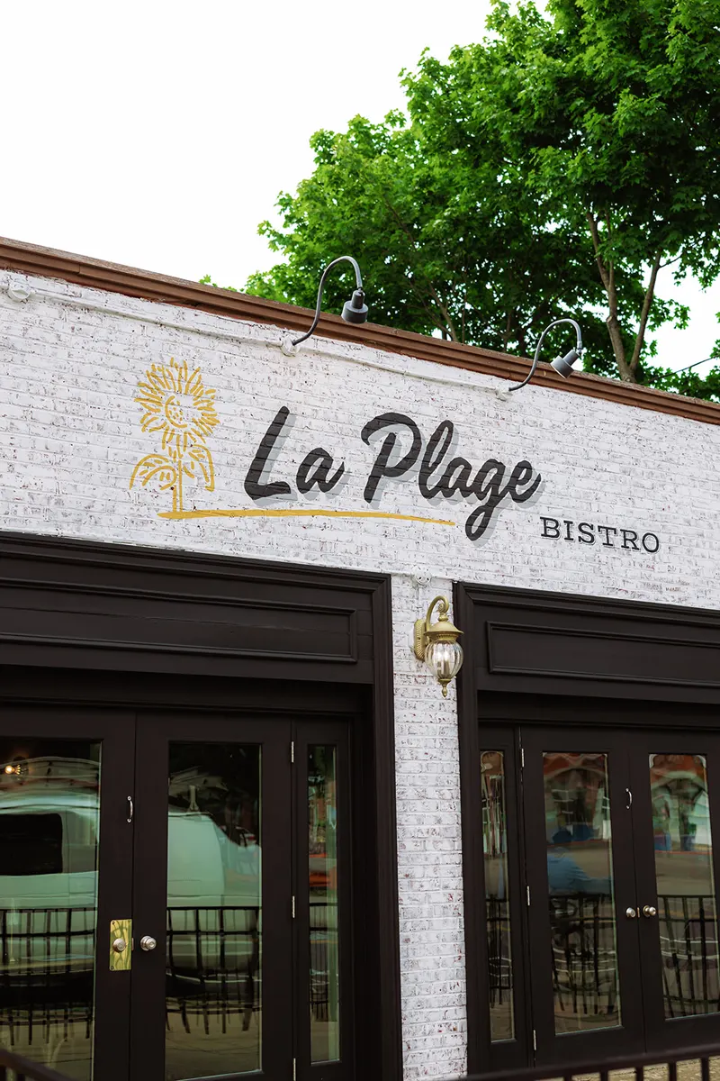 The exterior of La Plage Bistro, a charming French restaurant, features a white brick facade with the restaurant's name and logo above dark-framed glass doors.