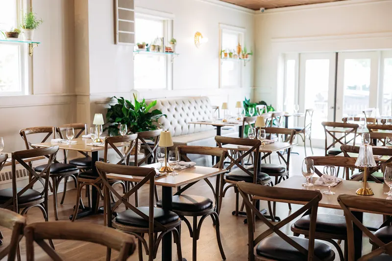 Bright, modern French restaurant dining area with wooden tables and chairs, set with glassware and napkins, surrounded by plants and natural light from windows.