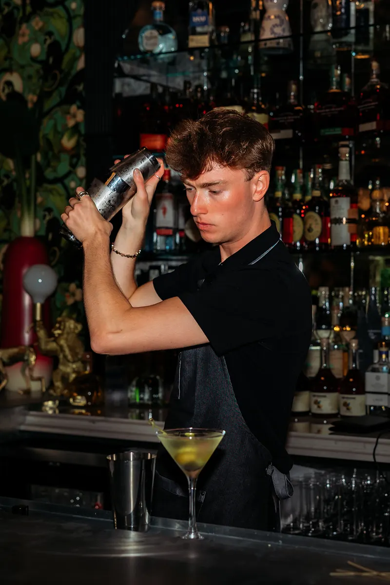A bartender in a black shirt and apron shakes a cocktail shaker behind the bar of a French restaurant, with a martini glass on the counter in front of him and bottles on shelves in the background.