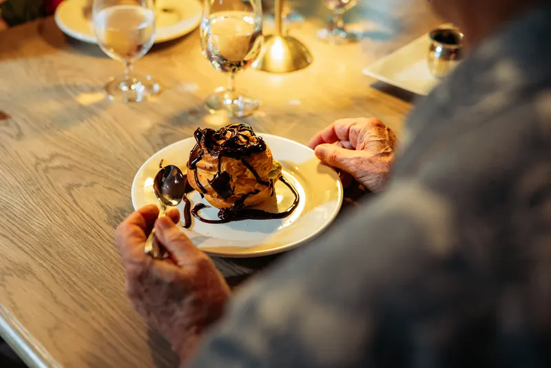 An elderly person holds a plate with a pastry dessert drizzled with chocolate sauce, seated at a wooden table set with glasses and plates in a cozy French restaurant.