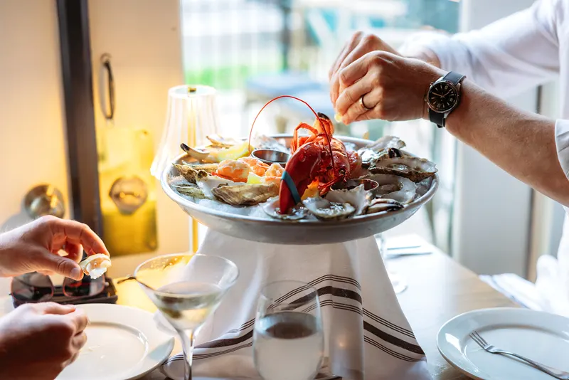 Two people share a seafood platter with oysters, clams, and lobster at a French restaurant table set with drinks and plates.