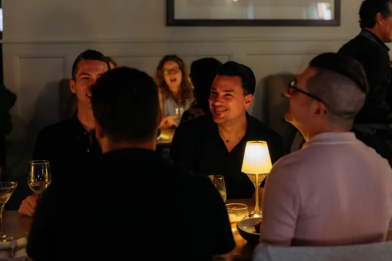 Four people are seated at a dimly lit French restaurant table, engaged in conversation, with drinks and a small table lamp in front of them. Other diners are visible in the background.