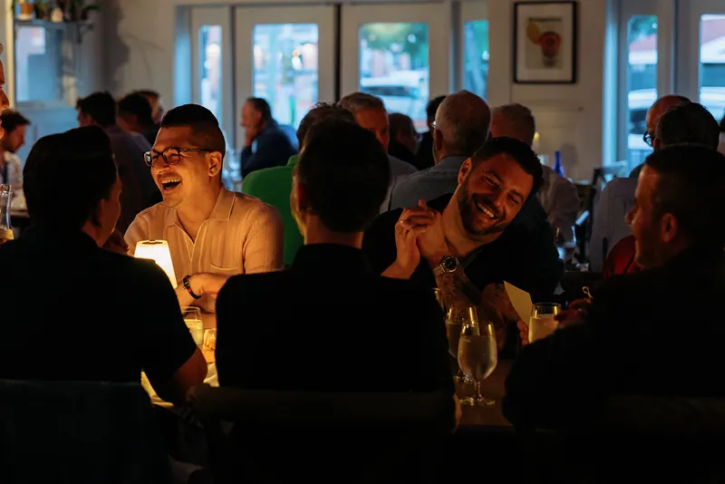 A group of people sit around a table at a dimly lit French restaurant, smiling and laughing while talking and enjoying drinks.