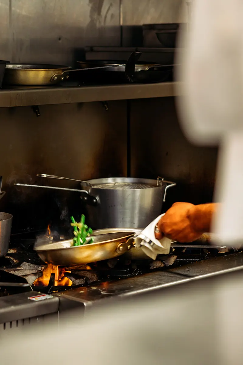A person holds a pan with vegetables over an open flame on a commercial kitchen stove in a bustling French restaurant, with pots and pans visible in the background.