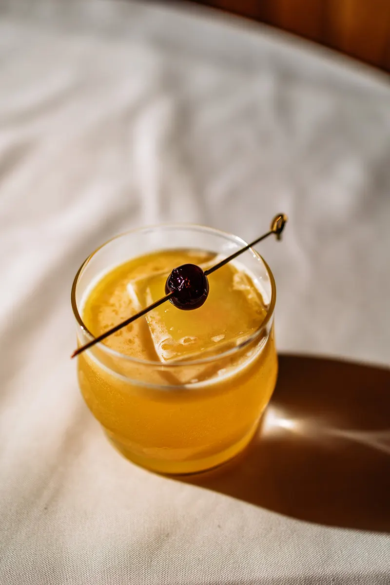 A glass filled with an orange-yellow cocktail over ice, garnished with a dark cherry on a cocktail pick, sits on a white tablecloth in a cozy French restaurant.