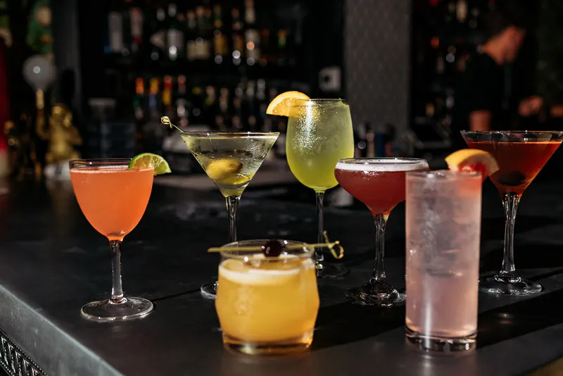 Seven assorted cocktails in various glassware are arranged on a bar counter in a French restaurant, with a blurred background of shelves stocked with bottles.