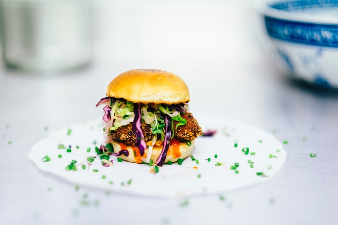 A small sandwich, reminiscent of a french restaurant classic, features a breaded patty, shredded cabbage slaw, and sauce on a white surface, garnished with chopped chives.