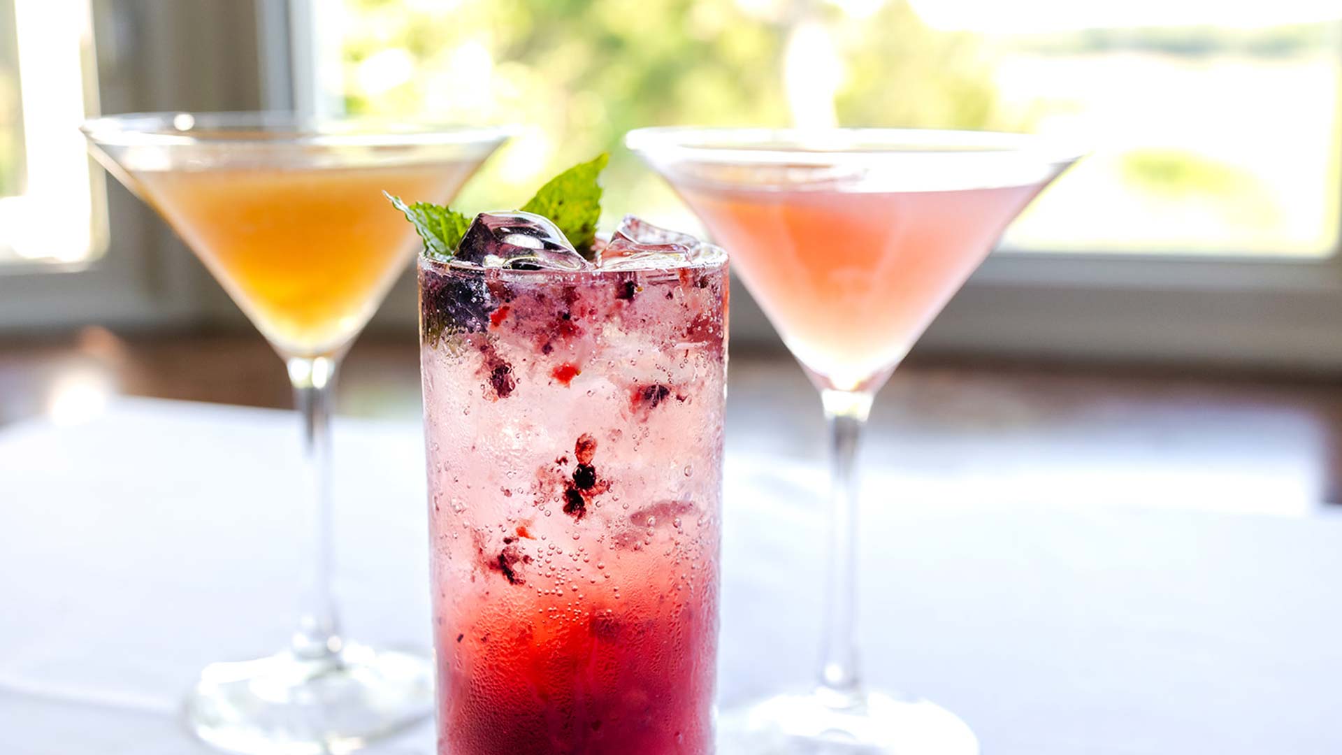 Three cocktails on a table: two in martini glasses with orange and pink hues, and one tall glass filled with a red, ice-filled drink garnished with mint.