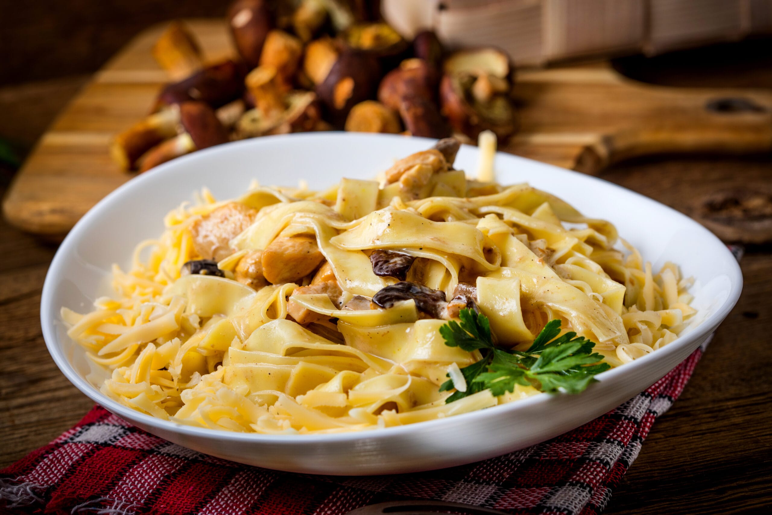 A white bowl filled with fettuccine pasta, chicken pieces, mushrooms, and grated cheese, garnished with parsley, evokes the charm of a French restaurant as it sits on a red checked cloth with mushrooms and a cutting board in the background.